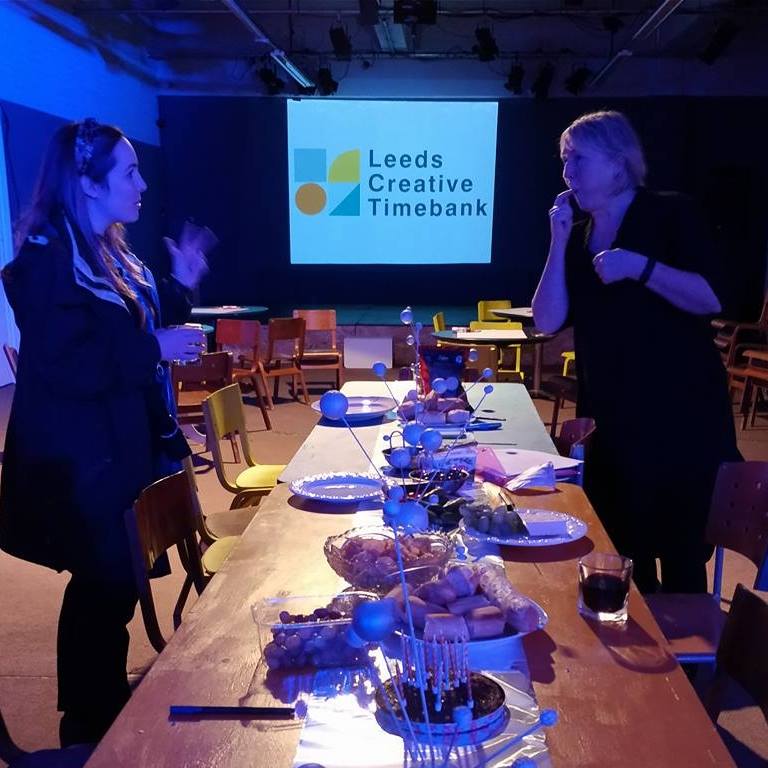 An event image from Winter Social and Design Hack showing Sue Ball discussing with a member over a table of food. The Timebank logo appears in the background.